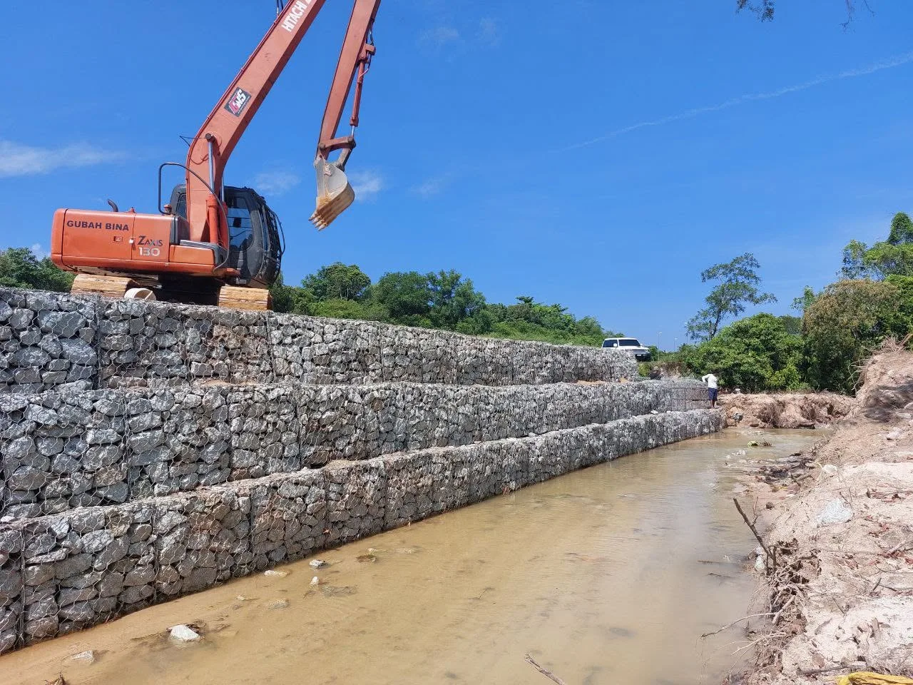 Tembok gabion bertingkat memanjang menyusuri laluan air, dengan jengkaut Gubah Bina berada di atas struktur.