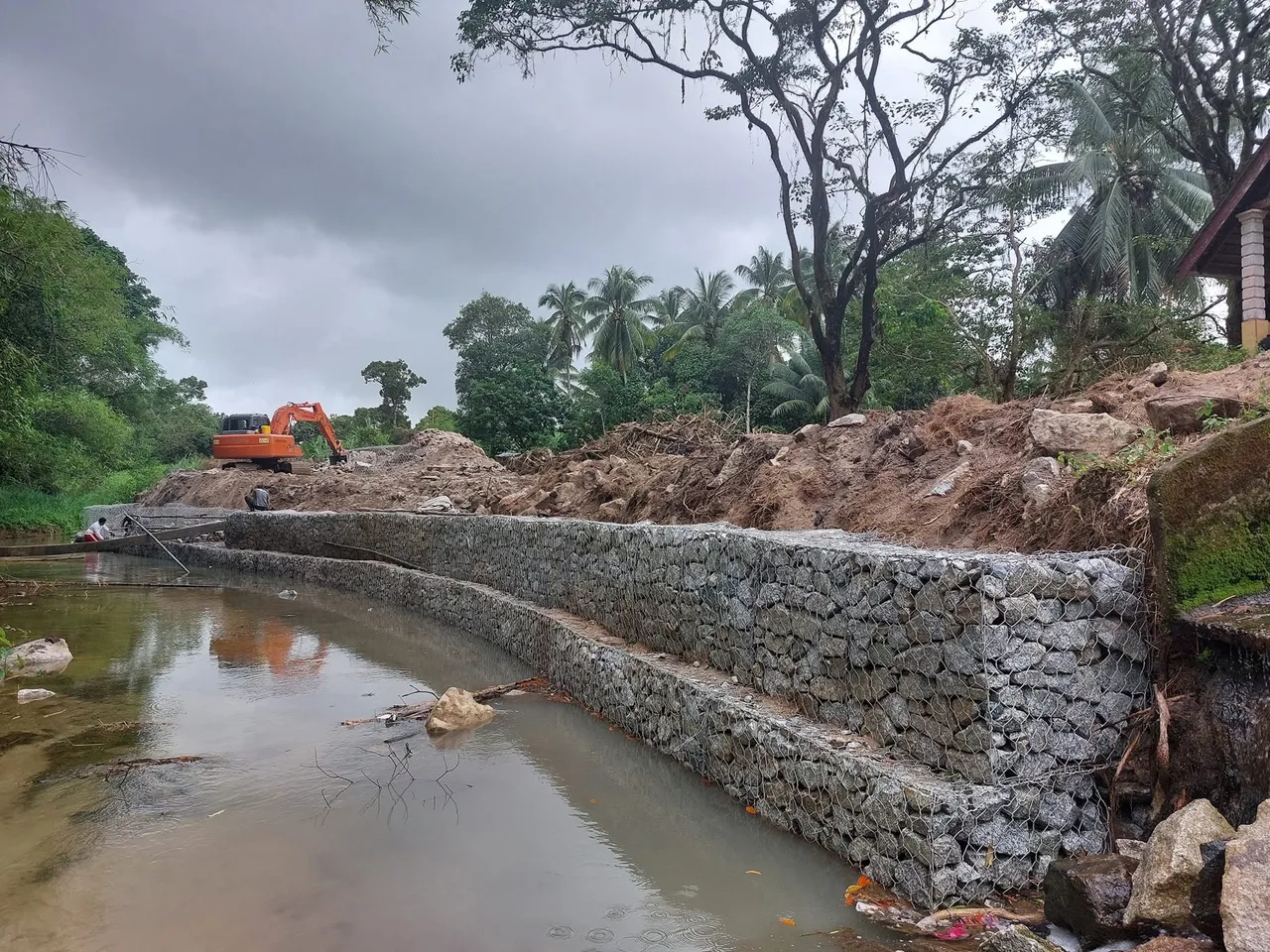 Kerja pemasangan gabion di tepi sungai sedang berjalan — sangkar disusun mengikut lengkung tebing dengan jengkaut di tapak.